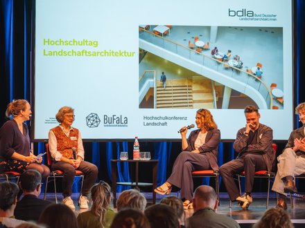 Podiumsdiskussion auf einer Bühne: fünf Personen sitzen auf roten Stühlen, eine spricht ins Mikrofon. Dahinter Leinwand mit „Hochschultag Landschaftsarchitektur“, Logos von Verbänden und Foto einer Treppenhalle.