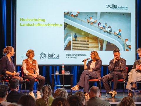 Podiumsdiskussion auf einer Bühne: fünf Personen sitzen auf roten Stühlen, eine spricht ins Mikrofon. Dahinter Leinwand mit „Hochschultag Landschaftsarchitektur“, Logos von Verbänden und Foto einer Treppenhalle.
