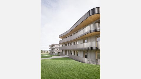 Seitliche Ansicht mehrerer Wohnhäuser mit gerundeten Balkonen, Holzfassade und großzügiger Grünfläche im Vordergrund.