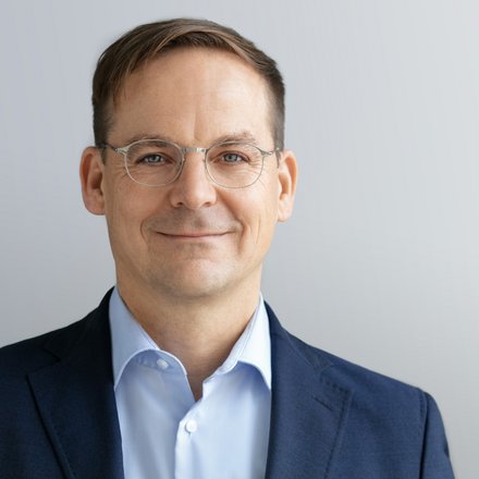 Kopf-und-Schulter-Porträt einer Person mit kurzem braunem Haar und dünner Metallbrille, in dunkelblauem Sakko und hellblauem Hemd, mit leichtem Lächeln vor hellgrauem, neutralem Hintergrund.