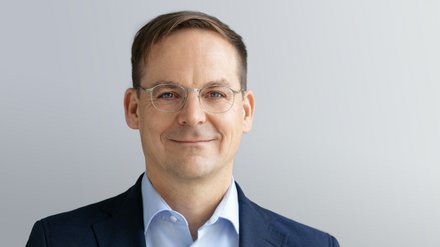 Kopf-und-Schulter-Porträt einer Person mit kurzem braunem Haar und dünner Metallbrille, in dunkelblauem Sakko und hellblauem Hemd, mit leichtem Lächeln vor hellgrauem, neutralem Hintergrund.