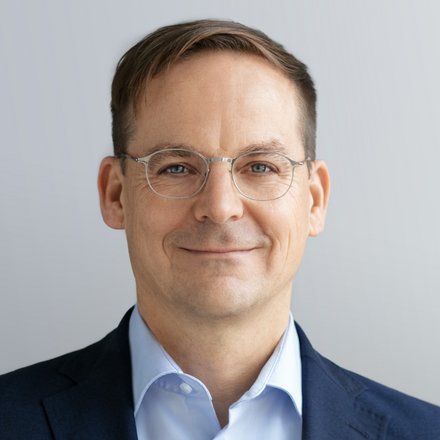 Kopf-und-Schulter-Porträt einer Person mit kurzem braunem Haar und dünner Metallbrille, in dunkelblauem Sakko und hellblauem Hemd, mit leichtem Lächeln vor hellgrauem, neutralem Hintergrund.