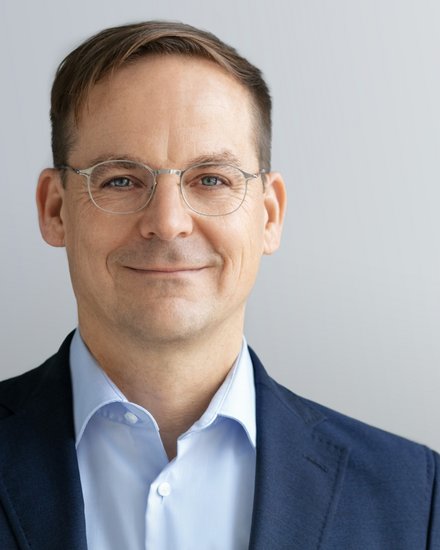 Kopf-und-Schulter-Porträt einer Person mit kurzem braunem Haar und dünner Metallbrille, in dunkelblauem Sakko und hellblauem Hemd, mit leichtem Lächeln vor hellgrauem, neutralem Hintergrund.