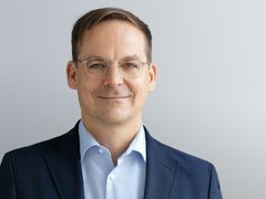 Kopf-und-Schulter-Porträt einer Person mit kurzem braunem Haar und dünner Metallbrille, in dunkelblauem Sakko und hellblauem Hemd, mit leichtem Lächeln vor hellgrauem, neutralem Hintergrund.