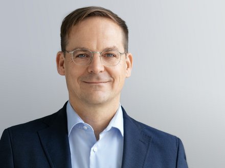 Kopf-und-Schulter-Porträt einer Person mit kurzem braunem Haar und dünner Metallbrille, in dunkelblauem Sakko und hellblauem Hemd, mit leichtem Lächeln vor hellgrauem, neutralem Hintergrund.