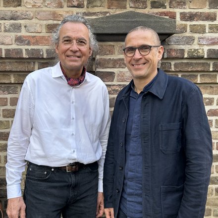 Zwei Männer vor einer Steinmauer lächeln in die Kamera.