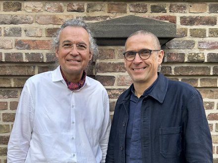 Zwei Männer vor einer Steinmauer lächeln in die Kamera.