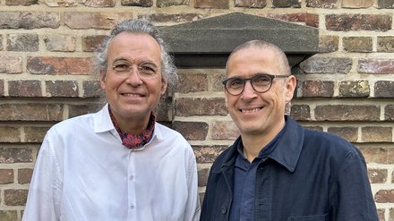 Zwei Männer vor einer Steinmauer lächeln in die Kamera.