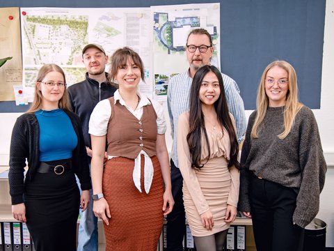 Gruppe aus Studierenden und Lehrendem vor Architekturplänen in einem Projektraum.