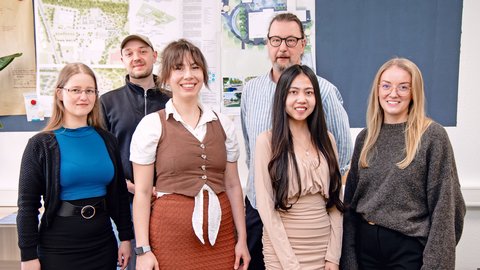 Gruppe aus Studierenden und Lehrendem vor Architekturplänen in einem Projektraum.