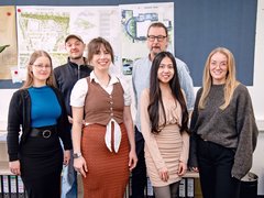 Gruppe aus Studierenden und Lehrendem vor Architekturplänen in einem Projektraum.
