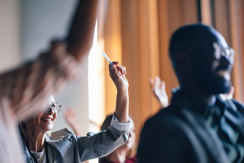 Mehrere Personen in einem Seminar oder Workshop heben die Hand; eine lächelnde Frau mit Brille im linken Bilddrittel hält einen Stift hoch, im rechten Vordergrund unscharfer Mann, warme Raumbeleuchtung im Hintergrund.