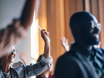 Mehrere Personen in einem Seminar oder Workshop heben die Hand; eine lächelnde Frau mit Brille im linken Bilddrittel hält einen Stift hoch, im rechten Vordergrund unscharfer Mann, warme Raumbeleuchtung im Hintergrund.
