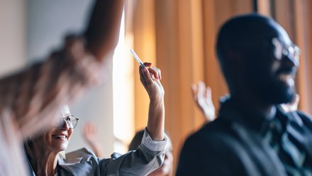 Mehrere Personen in einem Seminar oder Workshop heben die Hand; eine lächelnde Frau mit Brille im linken Bilddrittel hält einen Stift hoch, im rechten Vordergrund unscharfer Mann, warme Raumbeleuchtung im Hintergrund.