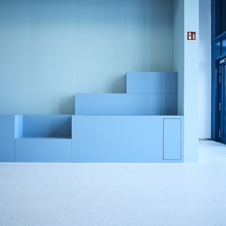 Ein minimalistischer Innenraum zeigt eine aus mehreren Stufen zusammengesetzte, geometrische Sitz- oder Podestlandschaft in verschiedenen Blautönen, die bündig in eine glatte Wand integriert ist, rechts öffnen bodentiefe Glasflächen den Raum zu einem hellen Flur oder Außenbereich, wodurch das Bild zeitgenössische Innenarchitektur mit klarer Formensprache darstellt und für flexible Nutzung, Aufenthaltsqualität und die Verbindung von Lernen, Begegnung und Raum im Kontext moderner Bildungs- oder Verwaltungsbauten steht.