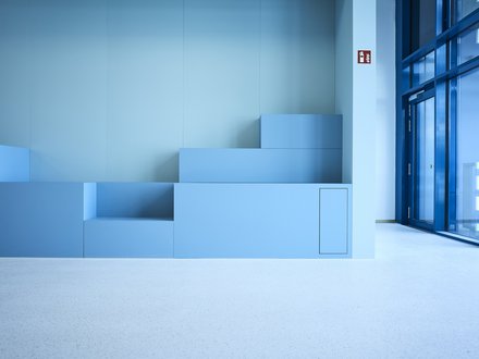 Ein minimalistischer Innenraum zeigt eine aus mehreren Stufen zusammengesetzte, geometrische Sitz- oder Podestlandschaft in verschiedenen Blautönen, die bündig in eine glatte Wand integriert ist, rechts öffnen bodentiefe Glasflächen den Raum zu einem hellen Flur oder Außenbereich, wodurch das Bild zeitgenössische Innenarchitektur mit klarer Formensprache darstellt und für flexible Nutzung, Aufenthaltsqualität und die Verbindung von Lernen, Begegnung und Raum im Kontext moderner Bildungs- oder Verwaltungsbauten steht.