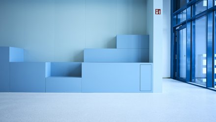 Ein minimalistischer Innenraum zeigt eine aus mehreren Stufen zusammengesetzte, geometrische Sitz- oder Podestlandschaft in verschiedenen Blautönen, die bündig in eine glatte Wand integriert ist, rechts öffnen bodentiefe Glasflächen den Raum zu einem hellen Flur oder Außenbereich, wodurch das Bild zeitgenössische Innenarchitektur mit klarer Formensprache darstellt und für flexible Nutzung, Aufenthaltsqualität und die Verbindung von Lernen, Begegnung und Raum im Kontext moderner Bildungs- oder Verwaltungsbauten steht.