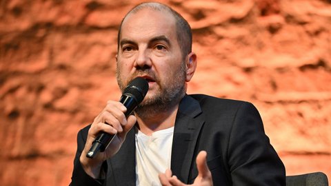 "Portrait eines Mannes mit dunklem Haar und Bart, der ein schwarzes Jackett über weißem Shirt trägt. Er hält ein Mikrofon und spricht engagiert. Seine Hände sind gestikulierend erhoben. Der Hintergrund zeigt unscharfe rote Backsteine einer historischen Mauer."