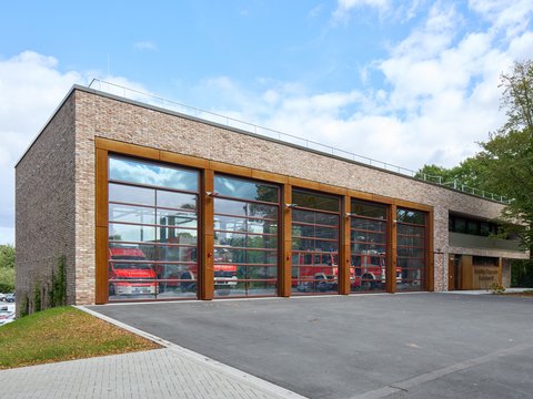 „Neubau eines Feuerwehrgerätehauses mit heller Klinkerfassade und fünf großen, verglasten Toren, die den Blick auf die roten Einsatzfahrzeuge freigeben.“
