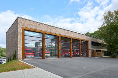„Neubau eines Feuerwehrgerätehauses mit heller Klinkerfassade und fünf großen, verglasten Toren, die den Blick auf die roten Einsatzfahrzeuge freigeben.“