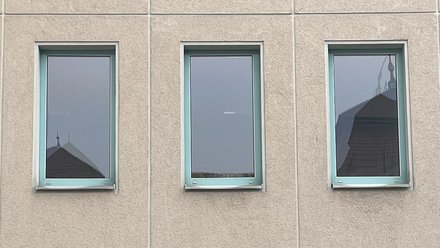 Drei schmale Fenster mit türkisfarbenen Rahmen in Putzfassade; im Glas spiegeln sich Dächer und Stadttürme.
