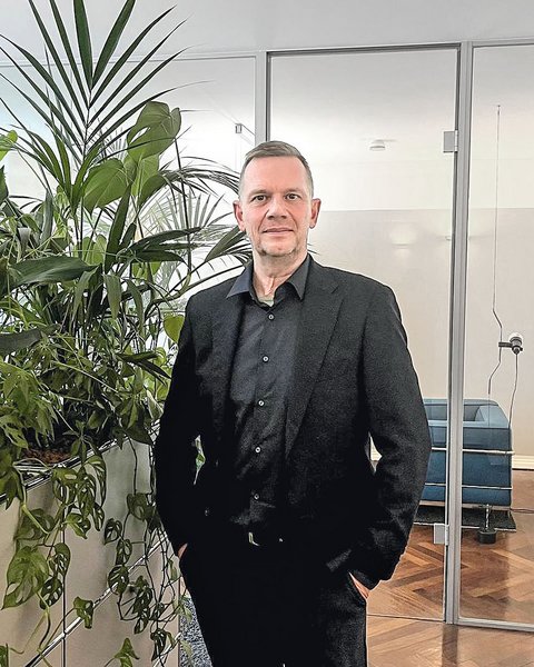Ein Mann mit kurzem grauem Haar und schwarzem Hemd steht in einem hellen Büro. Neben ihm befindet sich eine große Gruppe grüner Zimmerpflanzen, darunter eine Monstera und eine Palme. Im Hintergrund sind gläserne Türen, ein Sitzbereich mit blauem Sofa und weiße Büroschränke zu sehen.