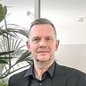 Ein Mann mit kurzem grauem Haar und schwarzem Hemd steht in einem hellen Büro. Neben ihm befindet sich eine große Gruppe grüner Zimmerpflanzen, darunter eine Monstera und eine Palme. Im Hintergrund sind gläserne Türen, ein Sitzbereich mit blauem Sofa und weiße Büroschränke zu sehen.