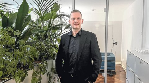 Ein Mann mit kurzem grauem Haar und schwarzem Hemd steht in einem hellen Büro. Neben ihm befindet sich eine große Gruppe grüner Zimmerpflanzen, darunter eine Monstera und eine Palme. Im Hintergrund sind gläserne Türen, ein Sitzbereich mit blauem Sofa und weiße Büroschränke zu sehen.