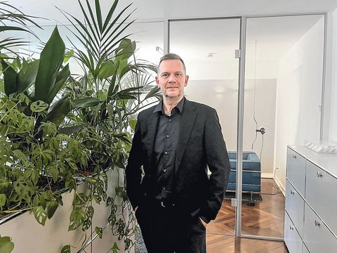 Ein Mann mit kurzem grauem Haar und schwarzem Hemd steht in einem hellen Büro. Neben ihm befindet sich eine große Gruppe grüner Zimmerpflanzen, darunter eine Monstera und eine Palme. Im Hintergrund sind gläserne Türen, ein Sitzbereich mit blauem Sofa und weiße Büroschränke zu sehen.