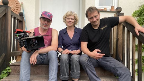 Drei Personen sitzen auf einer Treppe, links ein Mann mit Kamera und bunter Kappe, in der Mitte eine Frau mit kurzen lockigen Haaren, rechts ein Mann mit schwarzem T-Shirt und blauen Schuhen