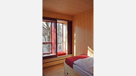 Modernes Schlafzimmer mit durchgehender Holzausstattung aus hellem Nadelholz. Großes Bett mit grauer Matratze und rotem Bettüberzug im Vordergrund. Breite Fensterbank mit roten Kissen an großem Fenster mit Ausblick auf Wald und Bäume. Dunkle Türrahmen. Holzdecke und Wände. Sonnenlicht fällt durch das Fenster und wirft Schatten auf die Holzwand. Minimalistisches, ruhiges Design