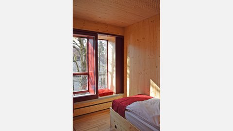Modernes Schlafzimmer mit durchgehender Holzausstattung aus hellem Nadelholz. Großes Bett mit grauer Matratze und rotem Bettüberzug im Vordergrund. Breite Fensterbank mit roten Kissen an großem Fenster mit Ausblick auf Wald und Bäume. Dunkle Türrahmen. Holzdecke und Wände. Sonnenlicht fällt durch das Fenster und wirft Schatten auf die Holzwand. Minimalistisches, ruhiges Design