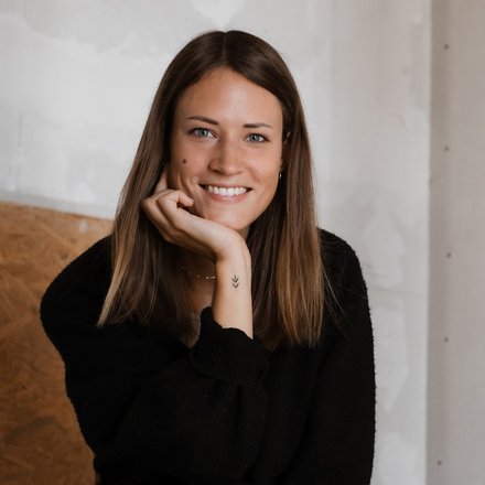 Porträt einer lächelnden Frau mit langen Haaren, sitzend in einem hellen Raum.