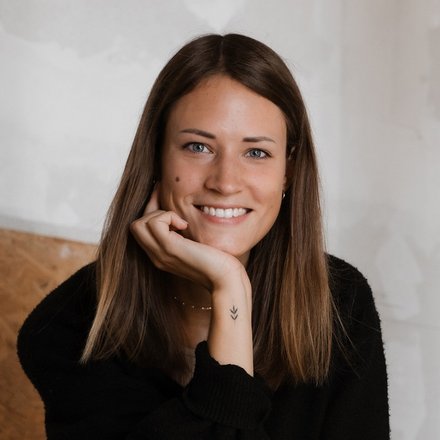 Porträt einer lächelnden Frau mit langen Haaren, sitzend in einem hellen Raum.