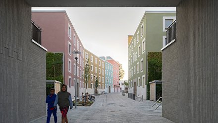 Schmale Wohnstraße zwischen modernen Gebäuden, zwei Personen gehen durch den Hofraum.