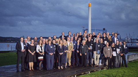 Breites Gruppenfoto einer Veranstaltung im Freien. Personen in mehreren Reihen, gemischt nach Alter und Geschlecht. Links und rechts Wiese, hinten Wasser mit Kaimauer, Kränen und Industrieanlagen; hoher weißer Mast mit orangefarbener Spitze. Dämmerlicht.