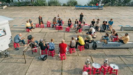 "Vogelansicht: Etwa 50 Personen verschiedenen Alters sitzen auf leuchtend roten Plastik-Hockern auf einem großen Platz. Mehrere sind im Kreis angeordnet, andere verteilt. Ein Hafengebiet mit Wasser und Industriegebäuden ist im Hintergrund sichtbar. Grüne Vegetation und Fahrräder rahmen die Szene ein."