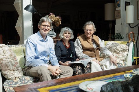 Drei ältere Personen sitzen auf einem Sofa in einem hellen Raum und lächeln in die Kamera