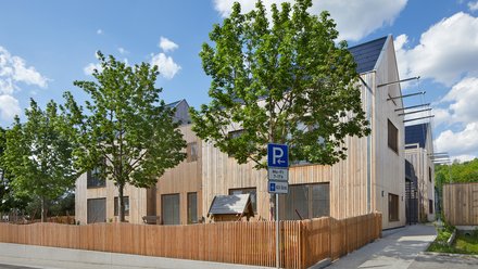 Straßenansicht: Kita mit Holzlattenzaun und jungen Bäumen; Parkplatzschild am Gehweg.