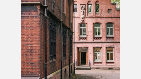 Historischer Backsteinbau mit Eingang, Rundbogenfenstern und Innenhof zwischen zwei Gebäudeflügeln.