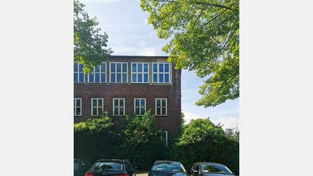 Backsteinfassade mit weißen Sprossenfenstern; davor geparkte Autos und dichte Begrünung, von Baumästen gerahmt.
