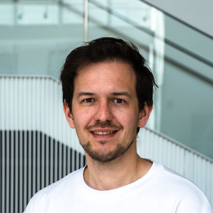  Porträt einer Person im weißen T‑Shirt, freundlich lächelnd, vor einer modernen Treppe mit Lamellenwand im Gebäude.