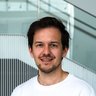  Porträt einer Person im weißen T‑Shirt, freundlich lächelnd, vor einer modernen Treppe mit Lamellenwand im Gebäude.