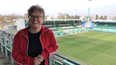 Eine Person in roter Jacke steht lächelnd auf der Tribüne eines Fußballstadions mit Blick auf das Spielfeld.