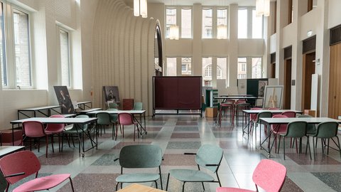 Großer heller Raum mit hohen Fenstern, mehreren Tischen und bunten Stühlen in Reihen und Gruppen angeordnet