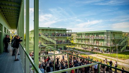 Außenansicht einer grünen, modularen Wohn- oder Campusanlage an sonnigem Tag: mehrere dreigeschossige Stahlbauten mit umlaufenden Laubengängen und Balkonen, vor den Gebäuden große Menschenmenge bei einer Versammlung oder Feier, auf Balkonen und Wegen viele Zuschauerinnen und Zuschauer, Perspektive von einem erhöhten Steg mit Geländer im Vordergrund.