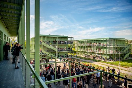 Außenansicht einer grünen, modularen Wohn- oder Campusanlage an sonnigem Tag: mehrere dreigeschossige Stahlbauten mit umlaufenden Laubengängen und Balkonen, vor den Gebäuden große Menschenmenge bei einer Versammlung oder Feier, auf Balkonen und Wegen viele Zuschauerinnen und Zuschauer, Perspektive von einem erhöhten Steg mit Geländer im Vordergrund.
