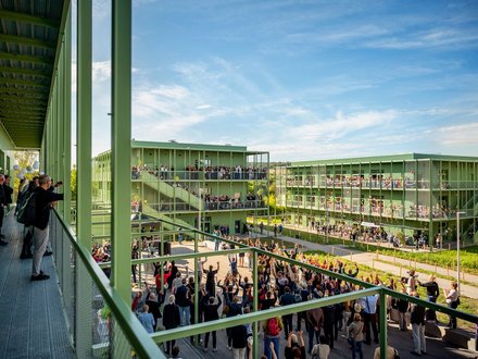 Außenansicht einer grünen, modularen Wohn- oder Campusanlage an sonnigem Tag: mehrere dreigeschossige Stahlbauten mit umlaufenden Laubengängen und Balkonen, vor den Gebäuden große Menschenmenge bei einer Versammlung oder Feier, auf Balkonen und Wegen viele Zuschauerinnen und Zuschauer, Perspektive von einem erhöhten Steg mit Geländer im Vordergrund.