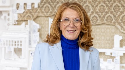 Halbnahes Porträt einer lächelnden, älteren Frau mit schulterlangem, rötlichem Haar und runder Brille; sie trägt ein hellblaues Sakko über einem kräftig blauen Rollkragenpullover und blickt direkt in die Kamera. Unscharfer Hintergrund mit weißen Architekturmodellen vor einer gemusterten Tapete.