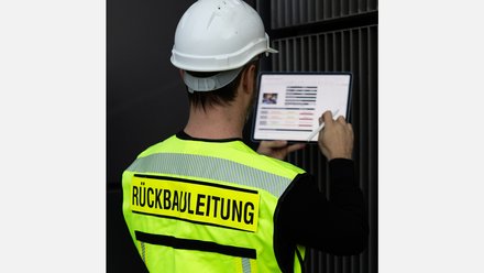 Person mit weißem Schutzhelm und gelber Weste „Rückbauleitung“ bedient ein Tablet auf einer Baustelle.